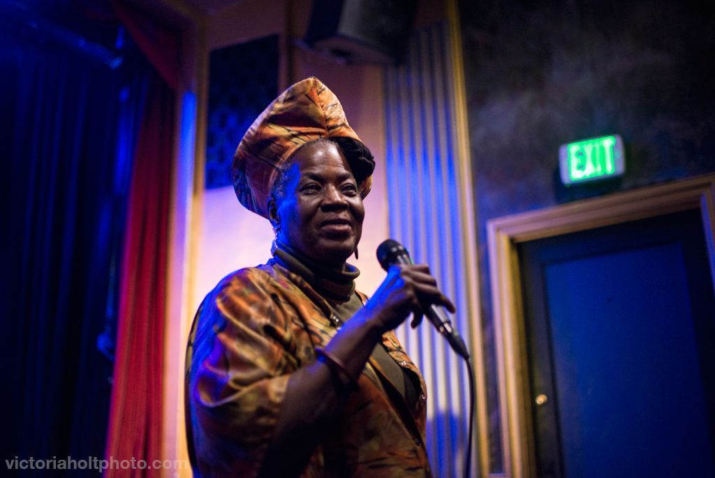 Storyteller Kibibi Monie speaking with a microphone and in cobalt lighting. Photo by Victoria Holt.